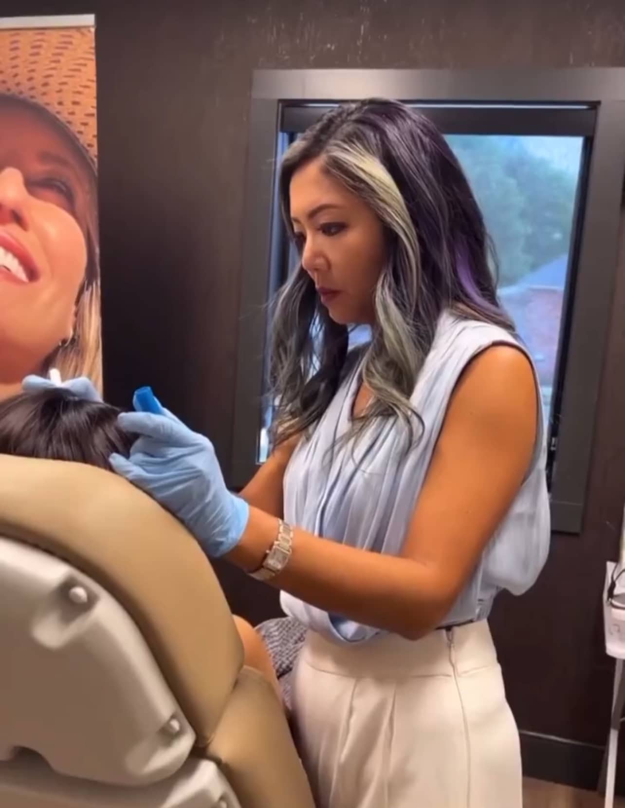 A woman with long hair and blue gloves administers an injection to a seated person in a room
