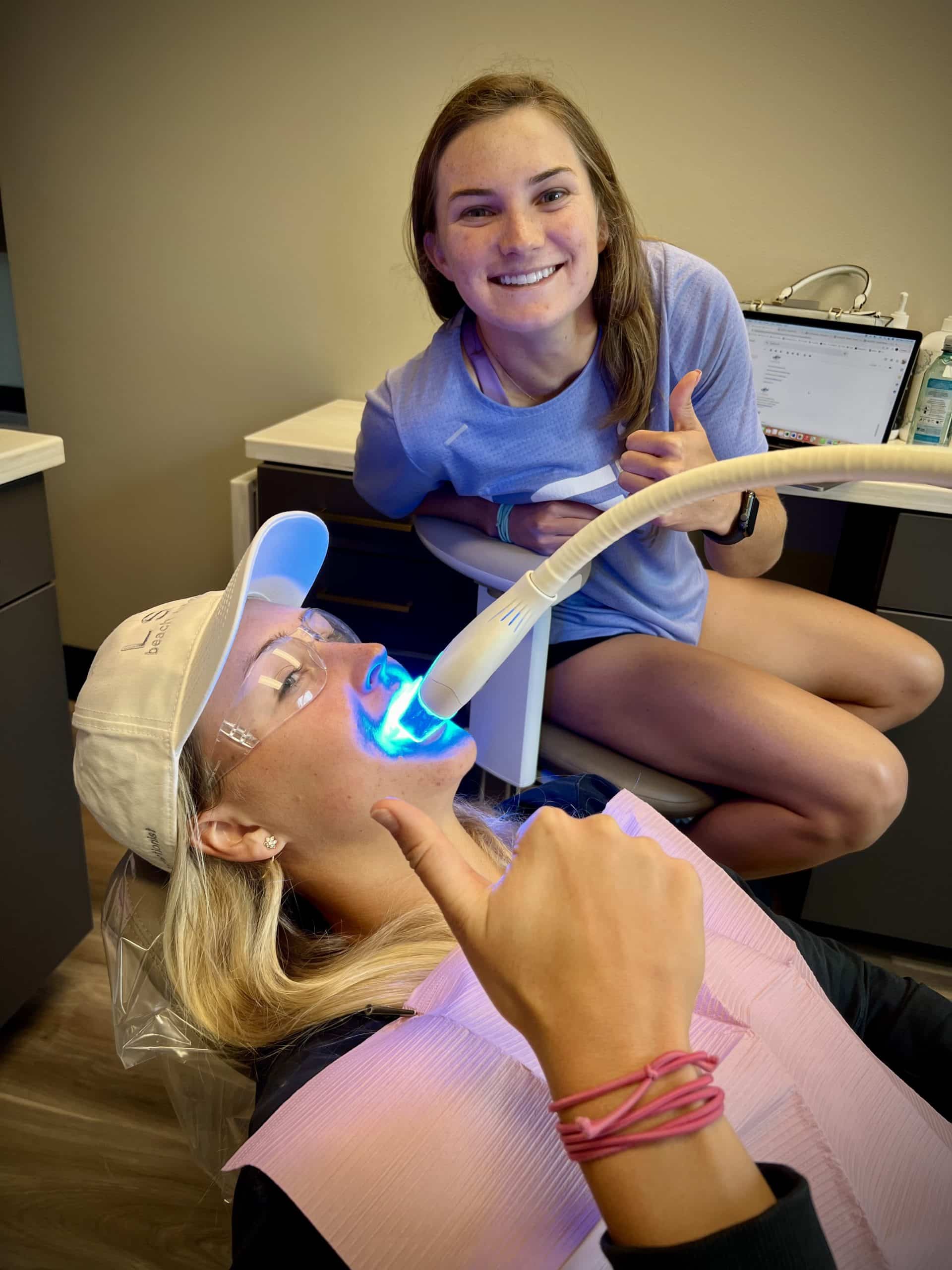 one person lying back with a whitening device in their mouth, and the other sitting beside