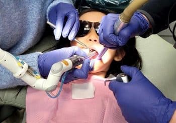 A child wearing sunglasses receives restorative dentistry treatment from multiple gloved hands using various dental instruments while lying in a dental chair.