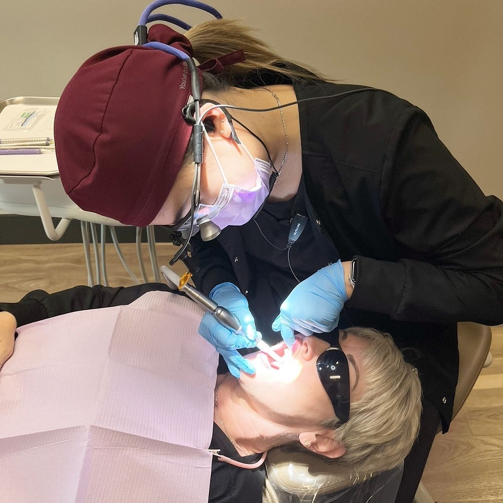 A dentist wearing a burgundy cap and magnifying loupes carefully performs a dental procedure to treat a knocked-out tooth on a patient lying back with a bib and sunglasses.