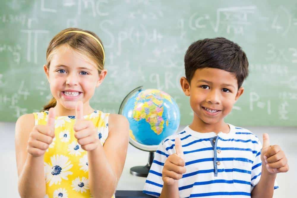 Two smiling children stand in front of a chalkboard and globe, giving thumbs up to the camera, confident after their tongue-tie treatment.