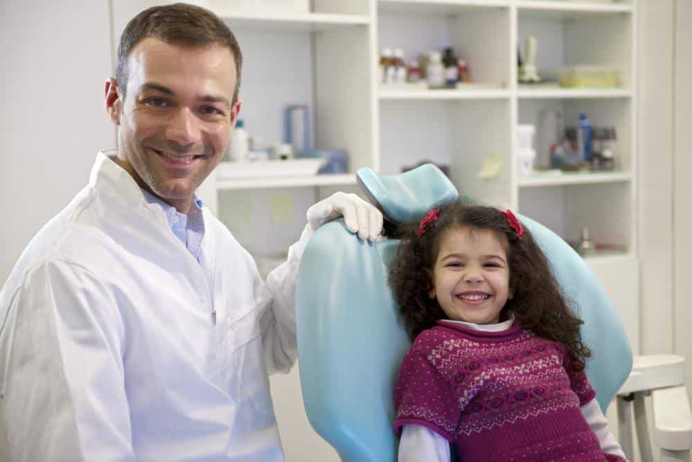 A dentist in a white coat smiles next to a young girl sitting in a dental chair, both looking at the camera in a dental clinic specializing in Tongue-Tie Treatment