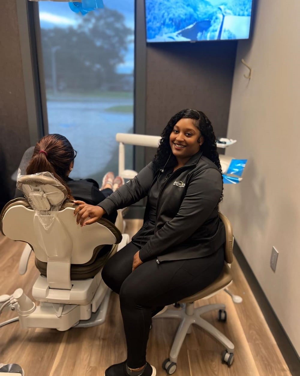 A dental hygienist sits beside a patient in a dental chair, holding the patient's arm. A monitor is on the wall and a window shows a rainy scene outside.