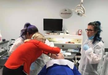Three people in lab coats assist a patient lying in a dental chair in a clinical setting with dental equipment visible.