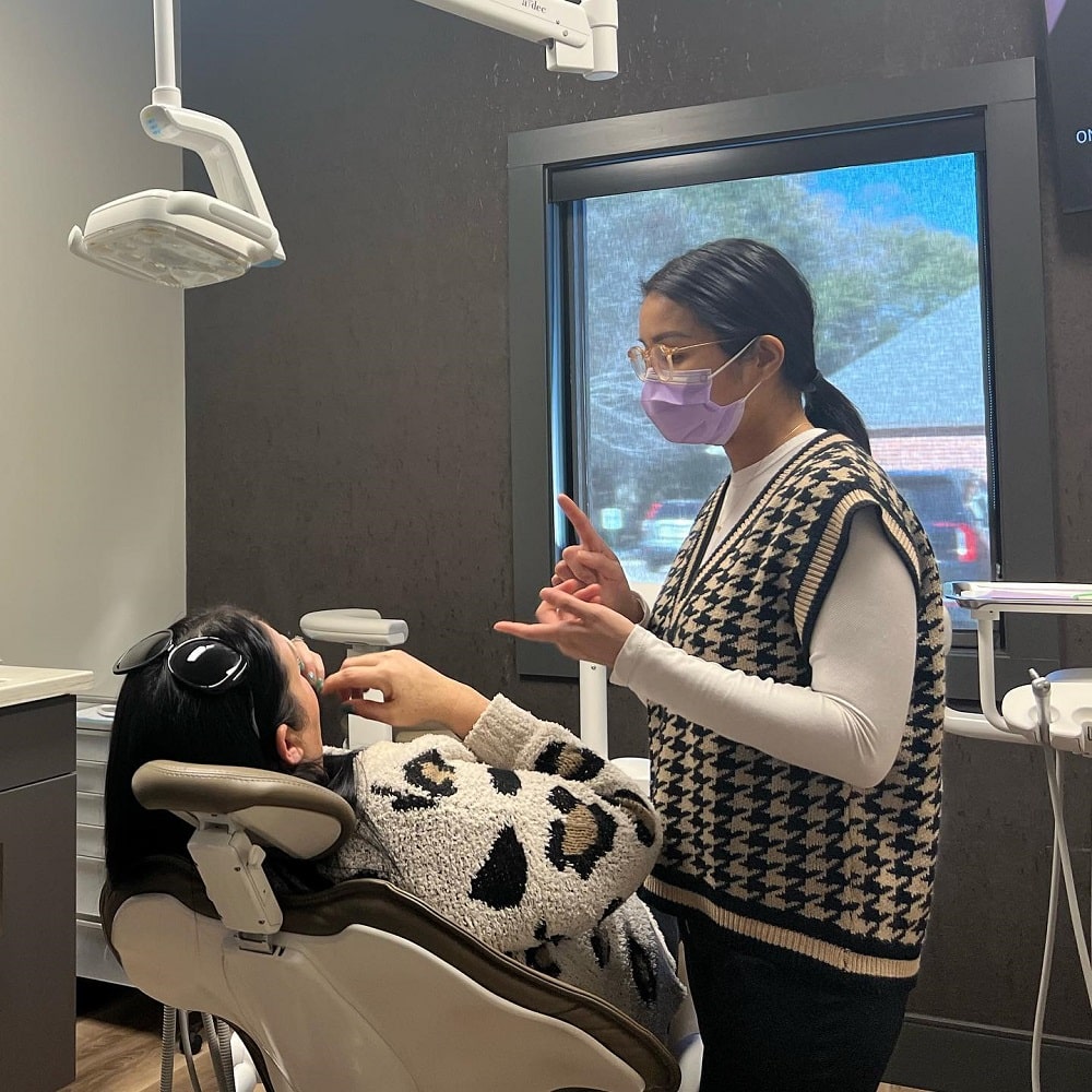 A dental professional wearing a mask explains a procedure to a patient sitting in a dental chair near a window.