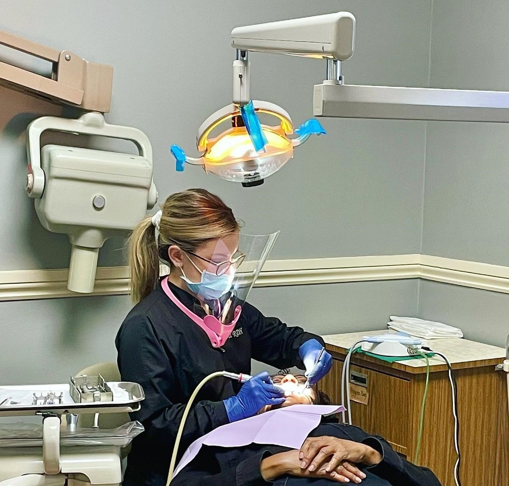 A dental professional wearing protective gear performs a dental procedure on a patient lying in a chair under an overhead exam light.