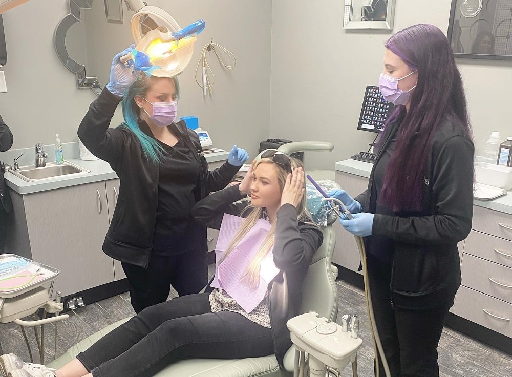 Two dental professionals prepare a female patient for a procedure in a dental clinic; one adjusts the light while the other holds dental tools.