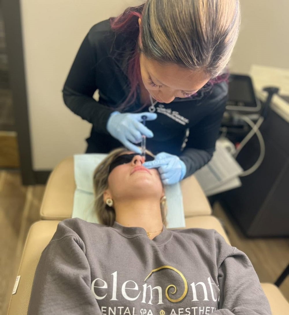A person lies on a treatment chair wearing sunglasses while a professional in gloves performs a cosmetic procedure on their face.