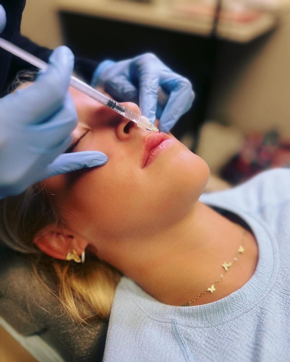 Person lying down receiving a cosmetic injection to the nose, with gloved hands holding a syringe and supporting the face.